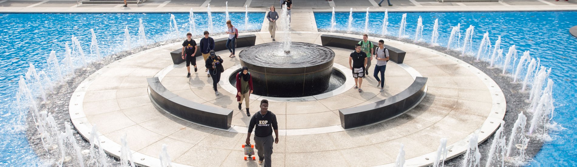 Several students walk along the bridge spanning the Podium Fountain. One student carries a skateboard and wears an EOP shirt.