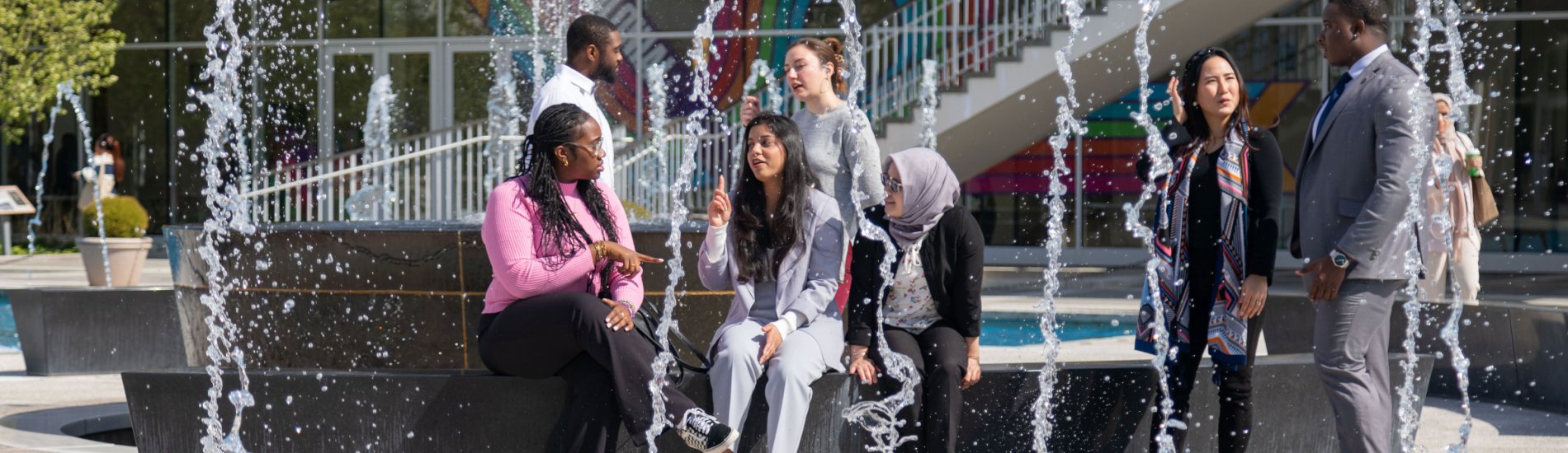 Seven student employees wearing business casual sit and stand around the main fountain on UAlbany's Uptown Campus.