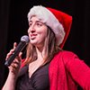 A female singer in red jacket and Santa hat sings into a mic