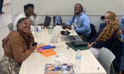 four people at a tablewith laptops and colorful flags smile at the camera