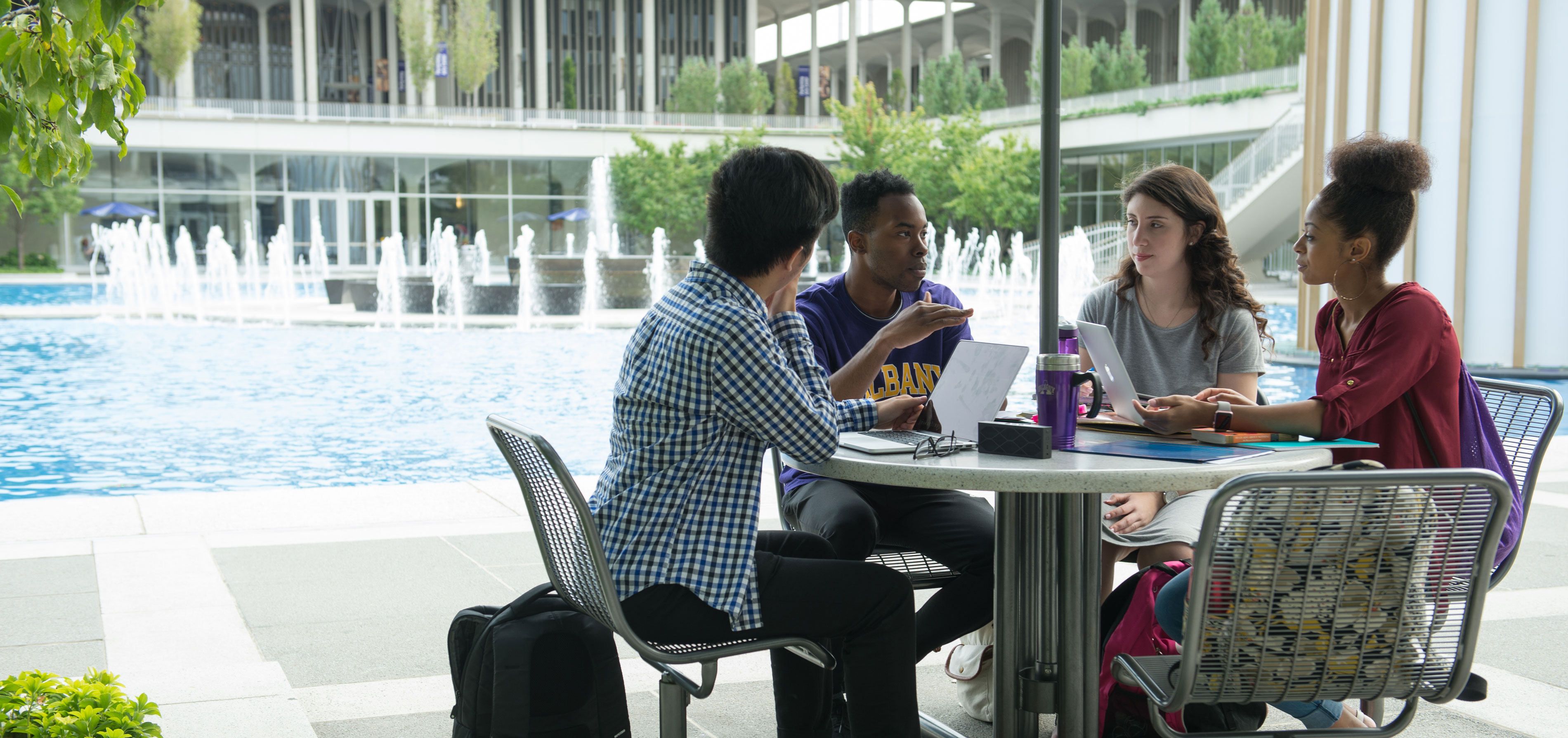 Four students sit at a table next to an outdoor fountain, with laptops and computers open in front of them as they converse.