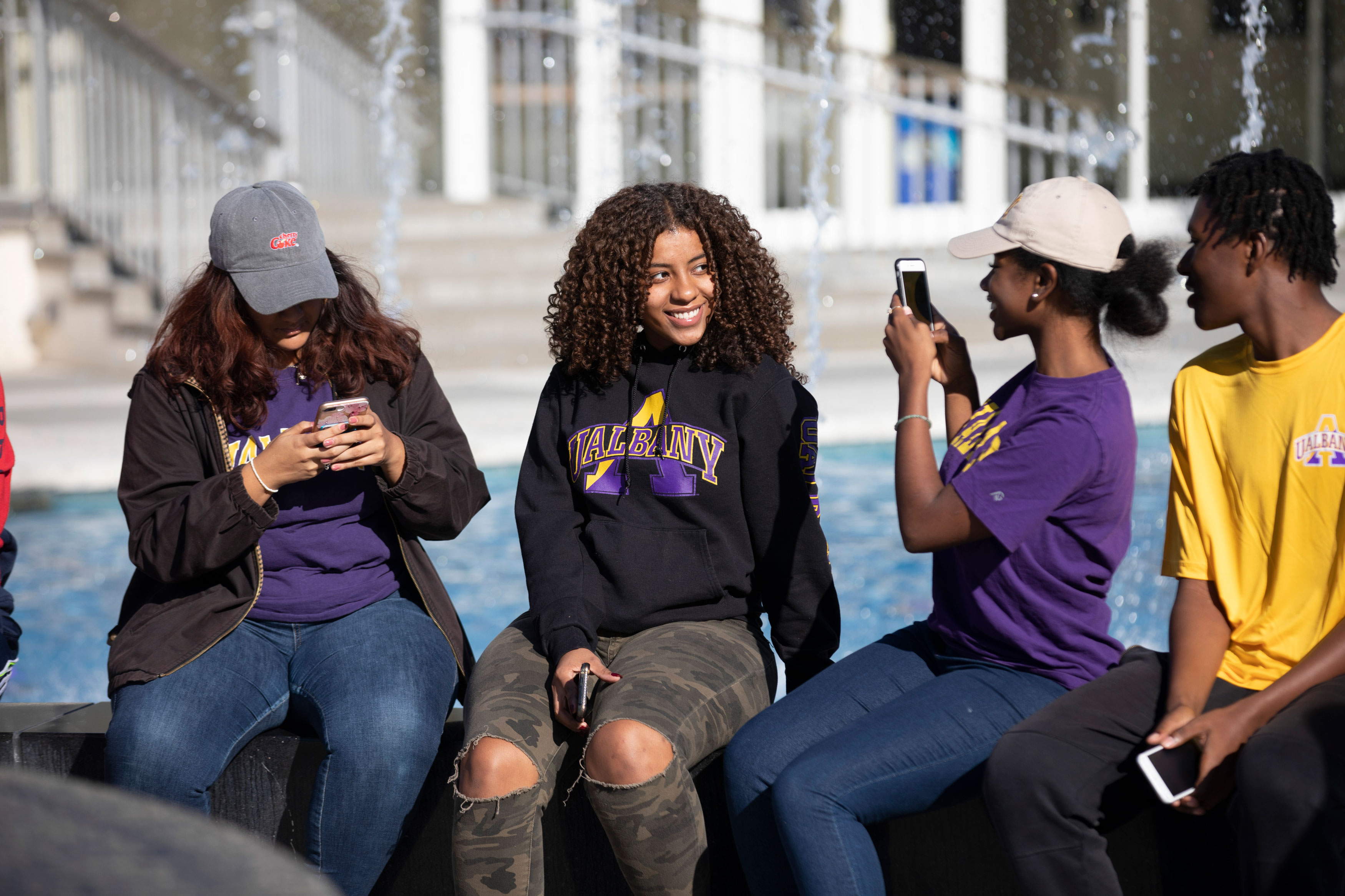 Four students sit on the edge of a fountain, one poses for a photo that the other is taking.