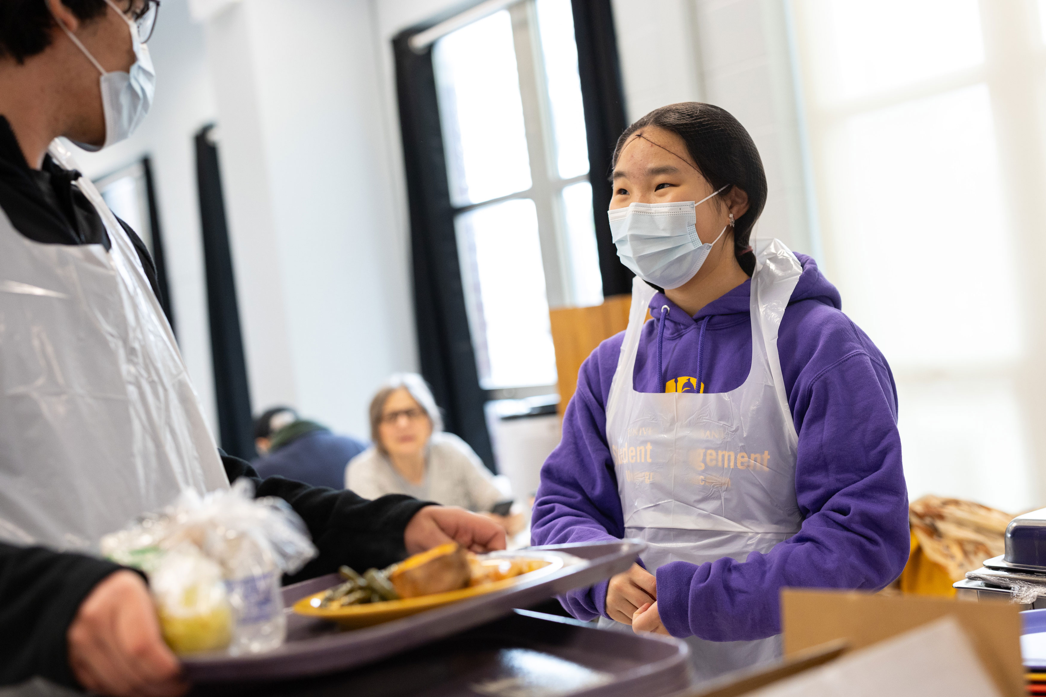 A student in a purple UAlbany sweatshirt, surgical mask, plastic apron smiles at another volunteer who is clearing trays at a soup kitchen.