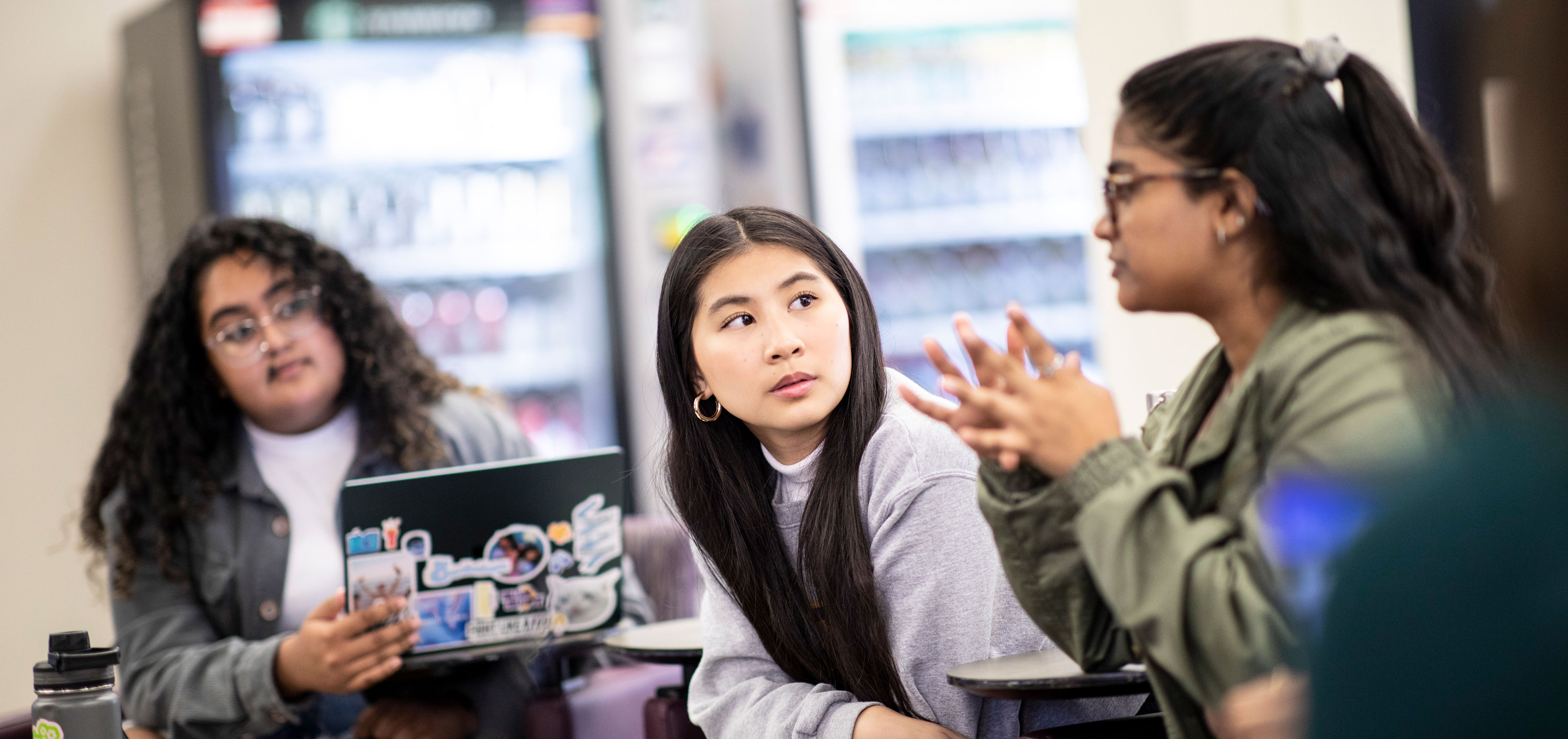 Two students sit inside the library listening as a third speaks and gestures.