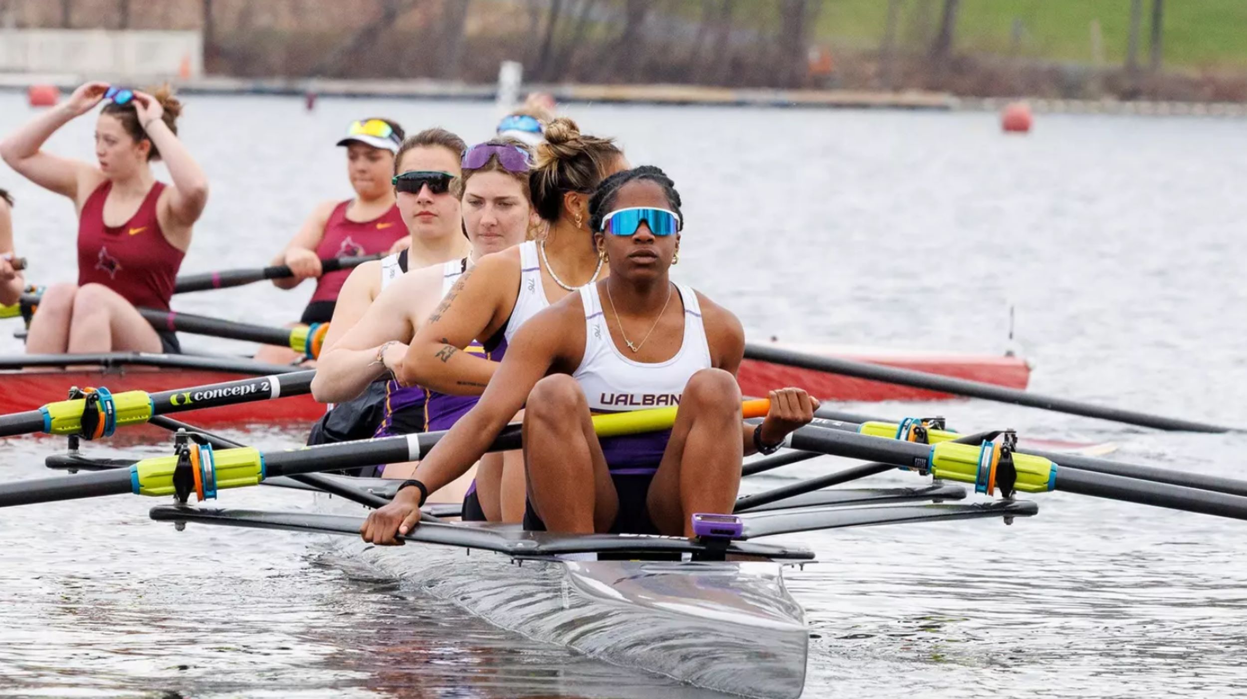 A group of four women rowers are in a sleak row boat on a calm body of water wearing jerseys with the letters UALBANY across the front.