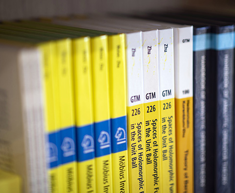 A row of mathematics books authored by Kehe Zhu sit on a bookshelf.