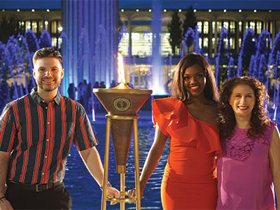 Two students and Alumni Association board president light torch at ceremony near main fountain.