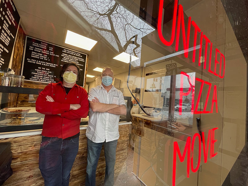 Shapiro and Bellucci stand inside store with full glass front and red lit sign that reas Untitled Pizza Movie