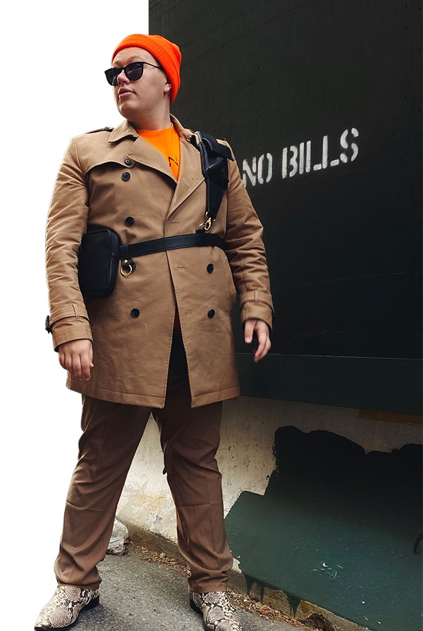 Russo poses wearing orange beanie, tan trench coat and matching pants