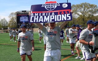 A man in a gray shirt and white shorts holds a sign that says America East Champions as his teammates walk behind him on a green athletic field during the day.